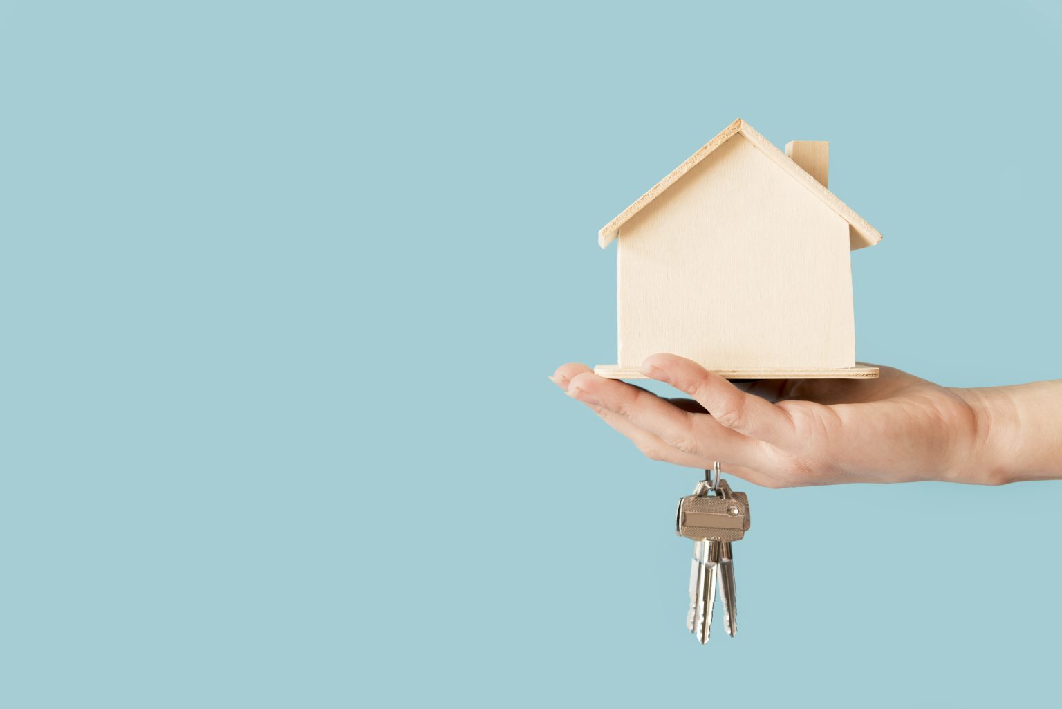 close up hand holding keys wooden house model against blue background